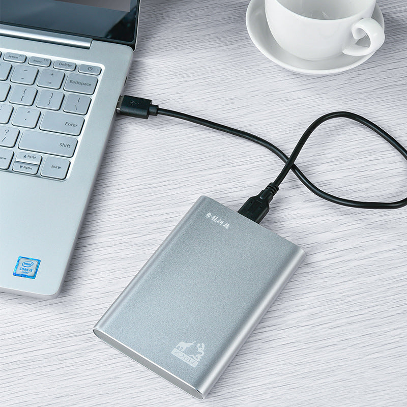 Silver metal mobile hard drive connected to a laptop via USB 3.0 cable on a white wooden desk next to a white coffee cup